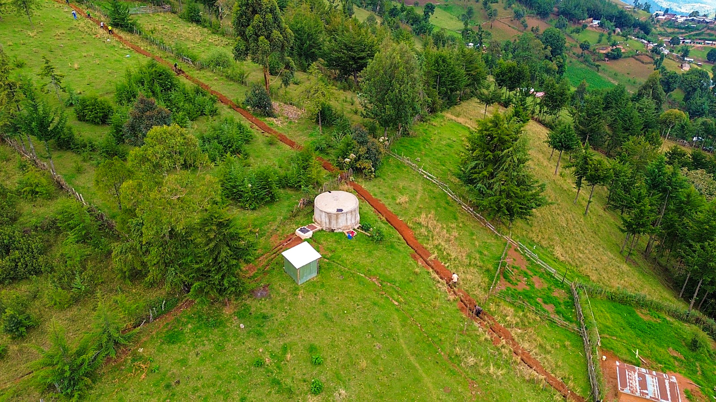 Aerial view of a hand-dug pipeline trench running down a green hillside to a water tank